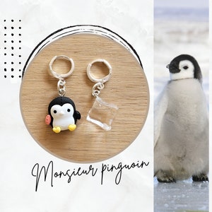 Può includere: Un paio di portachiavi argentati con un ciondolo a forma di pinguino bianco e nero e un ciondolo in acrilico trasparente. I portachiavi sono su una superficie di legno con la scritta "Monsieur pingouin".