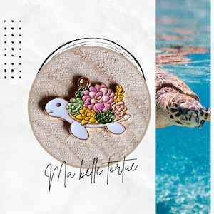 Może przedstawiać: Zawieszka w kształcie żółwia wykonana z białego metalu z pozłacanym obramowaniem. Żółw niesie na grzbiecie różnorodne kolorowe sukulenty. Zawieszka znajduje się na białym tle z napisem "Ma belle tortue" umieszczonym poniżej.