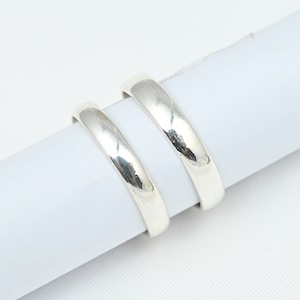 Puede incluir: Dos anillos plateados con una superficie lisa y pulida. Los anillos se colocan sobre un fondo blanco, mostrando su calidad reflectante. Las bandas tienen un diseño simple y clásico, adecuado para diversas ocasiones.