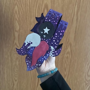 May include: A handmade, whimsical paper art piece featuring a stylized floral design. The artwork showcases a palette of deep purple, white, and red, with a star and cross embellishment. The design includes a dotted pattern and leaf-like shapes.