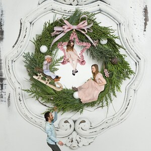 Könnte beinhalten: Ein skurriler Weihnachtskranz mit grüner Tannenbasis, verziert mit rosa Akzenten, weißen Ornamenten und einem dekorativen Stern. Der Kranz befindet sich in einem weißen, abgenutzten Rahmen, mit Figuren von Menschen und Kindern. Eine festliche und kreative Weihnachtsdekoration.