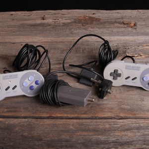 Puede incluir: Dos mandos grises de Super Nintendo con cables negros y un adaptador AV gris sobre una superficie de madera.