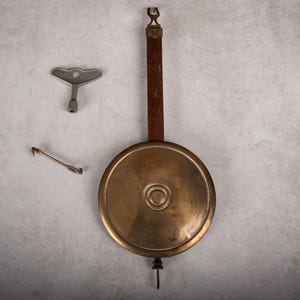 May include: A vintage clock pendulum with a brass face and wooden rod, accompanied by a winding key and a small adjustment tool. The pendulum is round and has a decorative design. The key and tool are silver-colored.