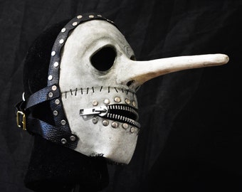 Könnte beinhalten: Eine weiße Totenkopfmaske mit einer langen, spitzen Nase, einem Reißverschluss für den Mund und einem schwarzen Lederband mit silbernen Nieten.