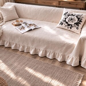 May include: A beige sofa covered with a ruffled slipcover and matching throw pillows. A decorative pillow with a black floral design rests on the sofa. A rug and a small side table with a lamp are in the background.
