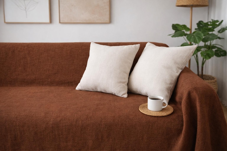 May include: A brown sofa with two off-white square pillows. A white mug with coffee sits on a small, round, woven coaster. The sofa is covered with a brown throw blanket. A houseplant and artwork are in the background.