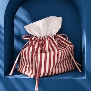 Może przedstawiać: Materiałowy pokrowiec na pudełko z chusteczkami w pionowe paski w kolorze ciemnoczerwonym i złamanej bieli. Prostokątny pokrowiec posiada u góry ściągacz z pasującymi tasiemkami, przez którego otwór wystaje biała chusteczka higieniczna.