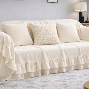 May include: A cream-colored sofa with a matching cover and three pillows. The cover has a ruffled skirt detail. The pillows include two smooth square pillows and one textured pillow. The sofa sits on a patterned rug.