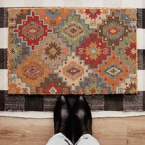 May include: A rectangular, patterned rug with a geometric design in shades of red, orange, blue, and brown. The rug is placed on a black and white checkered rug. Black leather boots and blue jeans are visible.