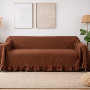 May include: A brown sofa cover with a ruffled edge, draped over a sofa. The cover is made of a textured fabric. The room also features a round side table, a floor lamp, and a potted plant. The overall theme is home decor.