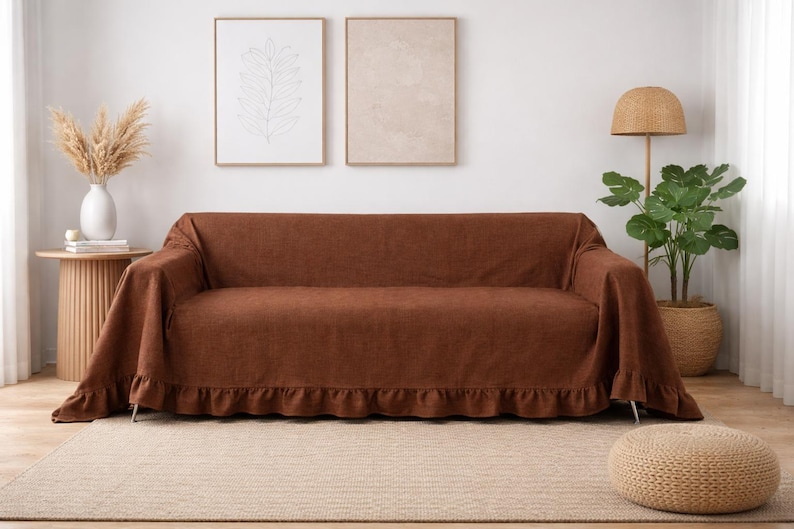 May include: A brown sofa cover with a ruffled edge, draped over a sofa. The cover is made of a textured fabric. The room also features a round side table, a floor lamp, and a potted plant. The overall theme is home decor.