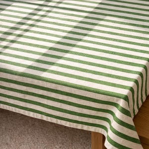 Puede incluir: Un mantel a rayas verdes y blancas. La tela tiene un aspecto natural y texturizado. Las rayas están espaciadas uniformemente y recorren horizontalmente la tela. El mantel está colocado sobre una mesa de madera, con la esquina de la tela visible.