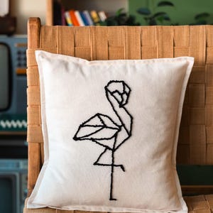 Könnte beinhalten: Quadratisches weißes Kissen mit einem schwarzen geometrischen Flamingo-Design. Das Kissen befindet sich auf einem gewebten Stuhl. Das Flamingo-Design besteht aus schwarzen Nähten. Ein Einrichtungsgegenstand.