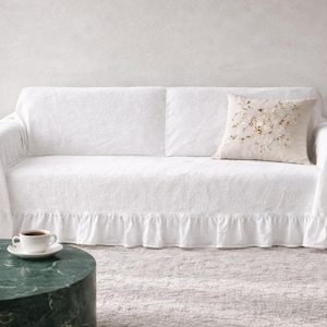 May include: A white sofa covered with a white linen slipcover featuring a ruffled skirt. A decorative pillow with gold and beige embellishments rests on the sofa. A cup of coffee sits on a white saucer on a green marble table.