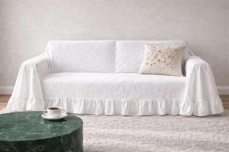 May include: A white sofa covered with a white linen slipcover featuring a ruffled skirt. A decorative pillow with gold and beige embellishments rests on the sofa. A cup of coffee sits on a white saucer on a green marble table.