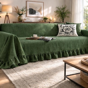 May include: A green sofa with a ruffled skirt is the focal point of a living room. A white pillow with a black rose design rests on the sofa. A wooden coffee table and plants complete the cozy scene.