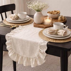 May include: A dining table set for a meal. The table is set with white plates, beige napkins, and gold silverware. A white vase holds white flowers. A lit candle and a bowl of snacks are also on the table. A beige table runner with a ruffled edge is in the center.
