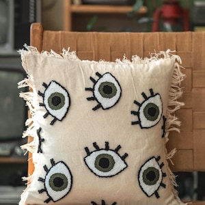 May include: Cream-colored square throw pillow with embroidered eye designs. The eyes have white, black, and olive green details. The pillow has a fringed edge and is placed on a woven chair.