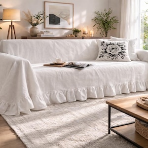 May include: A white linen sofa cover with a ruffled skirt. A black and white floral pillow rests on the sofa. The room is decorated with plants, a wooden coffee table, and a woven rug.