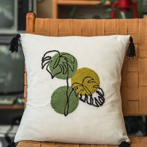 May include: Square white throw pillow with a green and yellow monstera leaf design. The leaves are embroidered with a textured, tufted appearance. Black tassels adorn each corner. The pillow is placed on a woven chair.