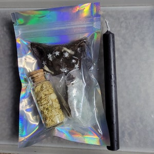 May include: A black ritual candle stands next to a clear holographic bag containing a dark brown, star-studded item, two small glass bottles with cork stoppers, and a white crystal-like substance. The bottles contain dried herbs.