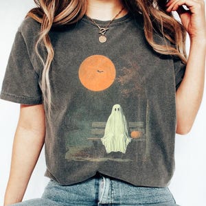 May include: Dark grey t-shirt with a Halloween graphic. The design shows a ghost on a park bench under an orange sun with a bat silhouette. A small pumpkin sits beside the ghost, evoking an autumnal scene.