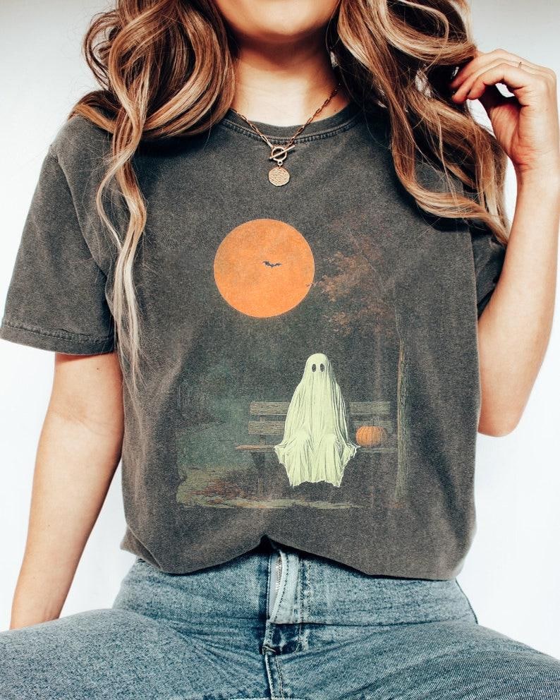 May include: Dark grey t-shirt with a Halloween graphic. The design shows a ghost on a park bench under an orange sun with a bat silhouette. A small pumpkin sits beside the ghost, evoking an autumnal scene.