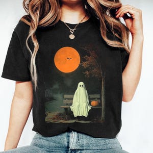 May include: Black t-shirt with a Halloween graphic. The design shows a ghost on a bench with a pumpkin, a large orange moon, and a tree. The shirt is dark and has a classic crew neck. The image evokes a spooky atmosphere.