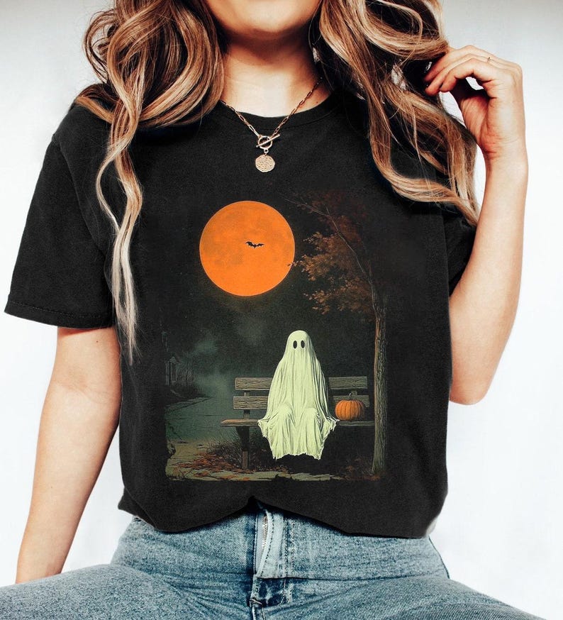 May include: Black t-shirt with a Halloween graphic. The design shows a ghost on a bench with a pumpkin, a large orange moon, and a tree. The shirt is dark and has a classic crew neck. The image evokes a spooky atmosphere.