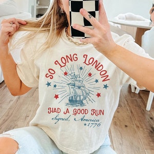 May include: A white t-shirt with a vintage-style graphic of a ship sailing away from a city skyline. The text "So Long London" is in a red circle around the ship, and the text "Had a Good Run Signed, America *1776" is below the ship.