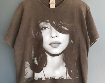 May include: A brown t-shirt with a black and white photo print of a woman with long dark hair. The woman is looking directly at the camera.