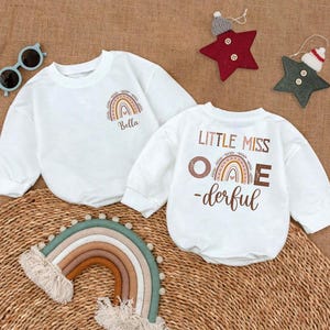 May include: Two white long-sleeved shirts with a rainbow design. One shirt says "Little Miss One-derful" and the other says "Bella" with a rainbow.