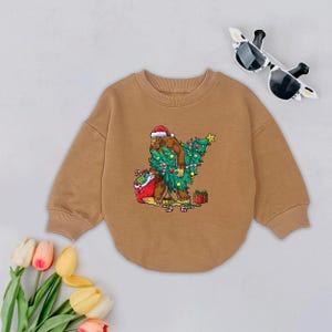 May include: A tan-colored sweatshirt featuring a graphic of a Bigfoot character wearing a Santa hat and carrying a Christmas tree. The design includes a gift bag, presents, and candy canes. Sunglasses are placed on the top right.