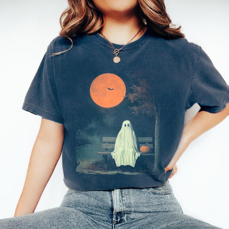 May include: Dark blue t-shirt with a Halloween graphic. The design shows a ghost on a bench under an orange moon, with a pumpkin. The shirt is a casual, short-sleeved style, suitable for autumn.