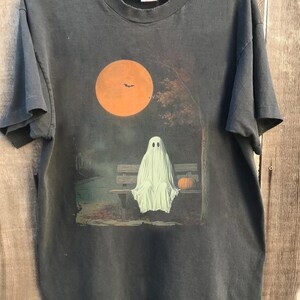 May include: Dark grey t-shirt with a Halloween design. A white ghost is seated on a park bench beneath a large orange moon with a bat silhouette. A small orange pumpkin sits beside the ghost. The shirt is on a wooden hanger.