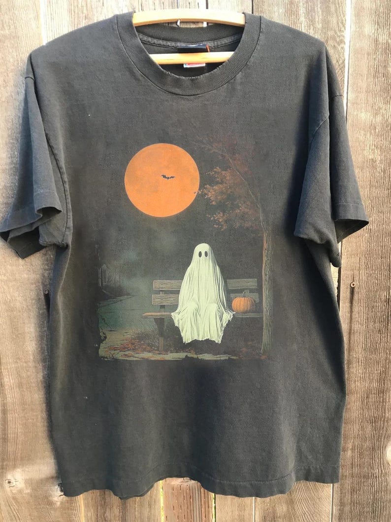 May include: Dark grey t-shirt with a Halloween design. A white ghost is seated on a park bench beneath a large orange moon with a bat silhouette. A small orange pumpkin sits beside the ghost. The shirt is on a wooden hanger.