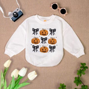 May include: White long-sleeved shirt with a Halloween theme. The shirt features a repeating pattern of orange pumpkins with black eyes and a black bow on each pumpkin.
