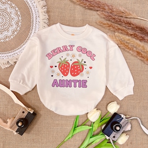 May include: A cream colored long-sleeved sweatshirt with the text "Berry Cool Auntie" in pink and purple letters. The text is surrounded by a graphic of two strawberries and flowers.