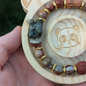 Può includere: Un braccialetto marrone e dorato con un ciondolo a forma di scoiattolo, un ciondolo a forma di fungo e varie perline di pietra. Il braccialetto è su una superficie di legno.
