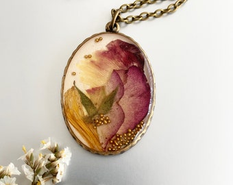 Op de afbeelding: Een gouden ketting met een ovale hanger met geperste bloemen en gouden glitter. De hanger is gevuld met helder hars en bevat een roze roos, gele bloemblaadjes en groene bladeren.