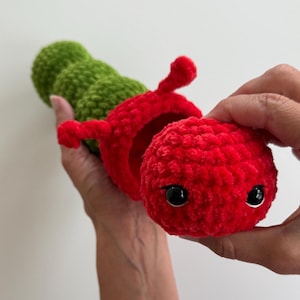 May include: A crocheted caterpillar toy in red and green. The caterpillar has a segmented body, with a red head featuring black eyes and antennae. The toy is made from a soft, textured yarn, suitable for children's play.