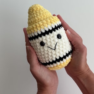 May include: A plush crochet toy shaped like a crayon, featuring a yellow pointed tip, a white cylindrical body, and black horizontal stripes. The toy has two small black plastic eyes and a stitched black V-shaped mouth on the front surface.