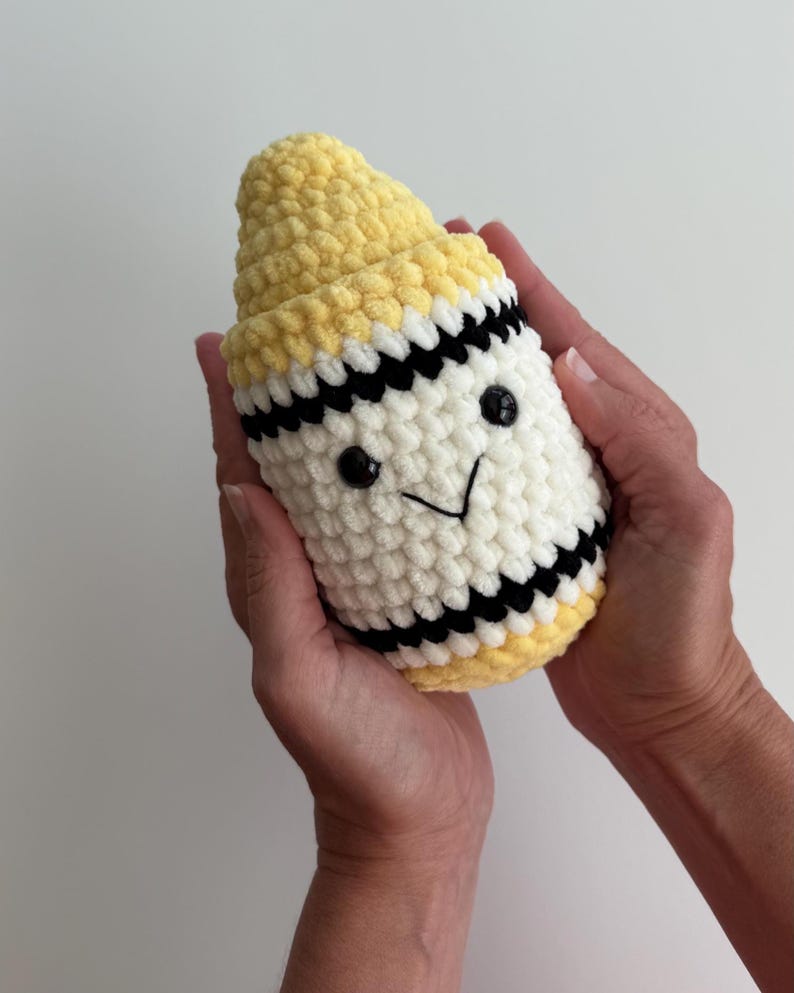 May include: A plush crochet toy shaped like a crayon, featuring a yellow pointed tip, a white cylindrical body, and black horizontal stripes. The toy has two small black plastic eyes and a stitched black V-shaped mouth on the front surface.