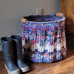 May include: A large, colorful crocheted basket with leather handles. The basket is filled with a variety of colors, including blue, purple, pink, white, and green. The basket is sitting on a wooden floor next to two black rain boots.