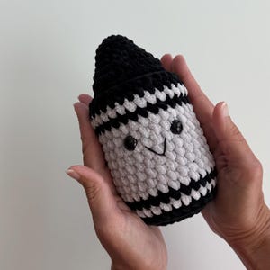 May include: A plush crochet toy shaped like a crayon, featuring a black pointed tip and a white body with horizontal black stripes. The toy has two small black plastic eyes and a stitched black V-shaped mouth, creating a simple facial expression.