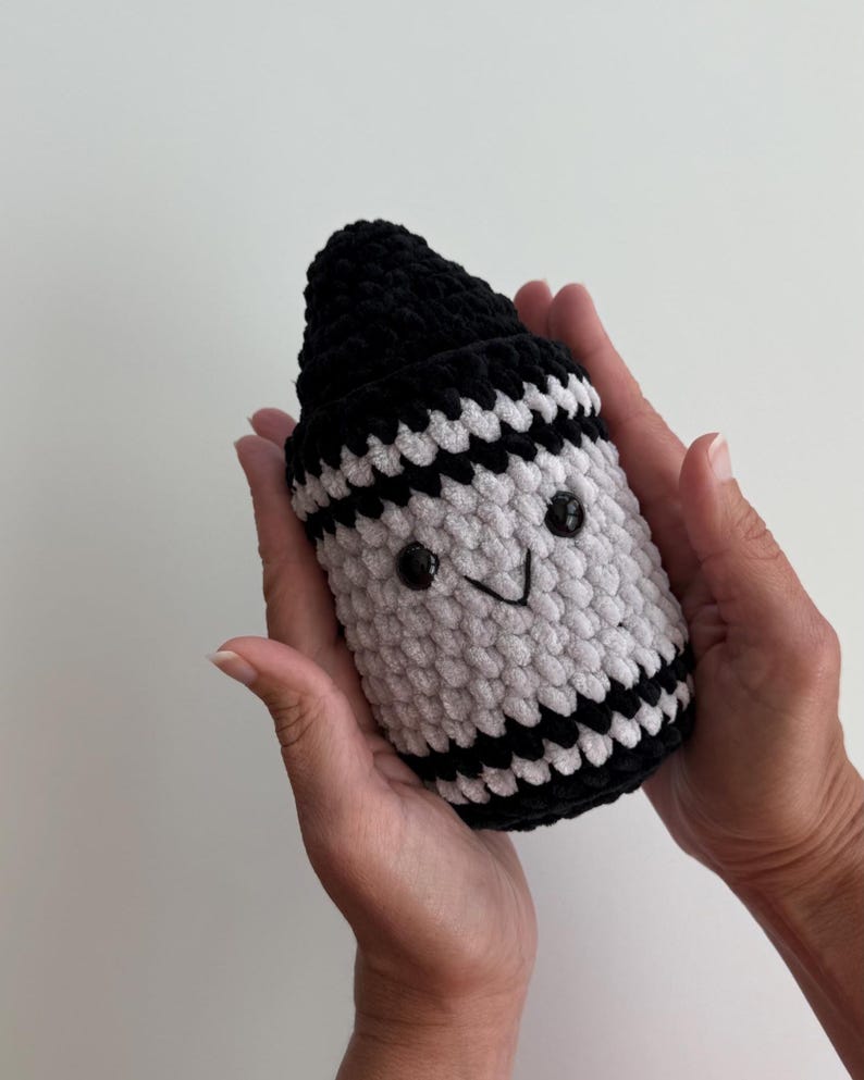 May include: A plush crochet toy shaped like a crayon, featuring a black pointed tip and a white body with horizontal black stripes. The toy has two small black plastic eyes and a stitched black V-shaped mouth, creating a simple facial expression.