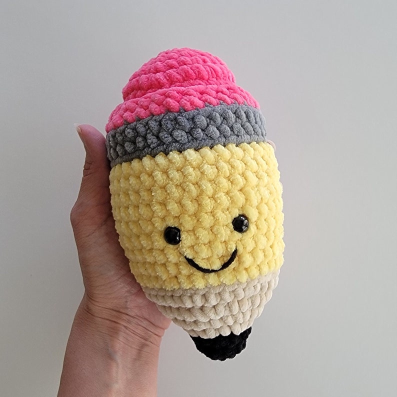 May include: A crocheted pencil plush toy with a pink eraser, yellow body, and a smiling face.