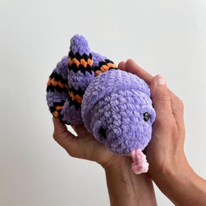 May include: A plush snake toy crafted from soft purple yarn, featuring a coiled body with alternating black and orange stripes. The stuffed animal has small black plastic eyes and a protruding light pink yarn tongue extending from its rounded head.