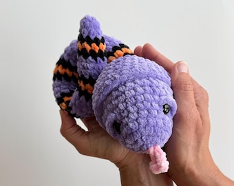May include: A plush snake toy crafted from soft purple yarn, featuring a coiled body with alternating black and orange stripes. The stuffed animal has small black plastic eyes and a protruding light pink yarn tongue extending from its rounded head.
