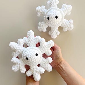 May include: Two white crocheted snowflake plushies with soft, textured yarn and small, round, black plastic eyes. Each snowflake features a central circular body with six radiating arms, each ending in a branched, crystalline-like point.